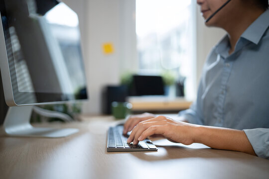 Young Asian man wearing a headset, typing on a keyboard at he desk, providing friendly call center and tech support in a modern home office environment