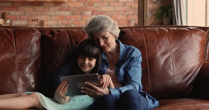 Grandmother and granddaughter spending time together on couch using digital tablet, watching video or educational cartoons online. Intergenerational family connection, modern technology use at home