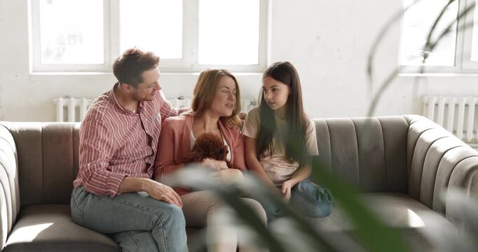 Smiling family spending time with toy poodle dog, resting seated on sofa in living room, talking, enjoying pastime with child daughter and cute domestic animal at home. Communication, love, pet care