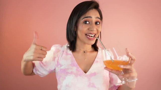 Close up portrait of happy smiling pretty young Indian woman girl hold glass enjoying delicious orange juice looking at camera showing thumb up, posing against light pink studio background.
