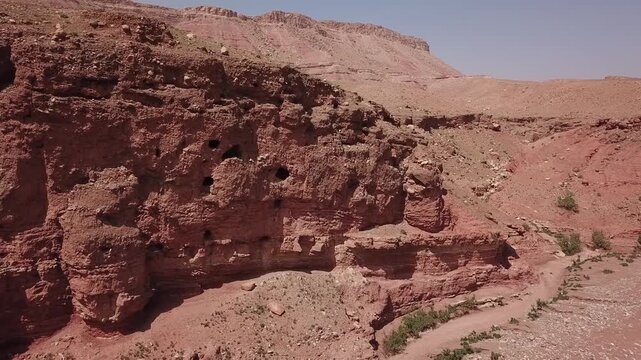 Drone shot of prehistoric cave dwellings carved into a sheer red sandstone cliff face within a rugged Moroccan canyon landscape.