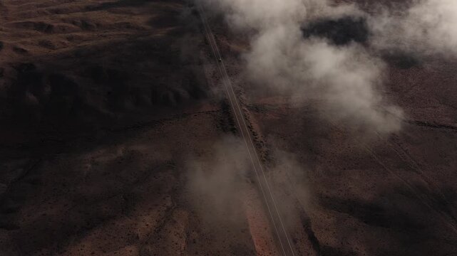 This drone perspective captures a vehicle traveling through a desolate and misty badlands environment. The atmospheric clouds and rugged desert terrain create a sense of mystery and isolation.