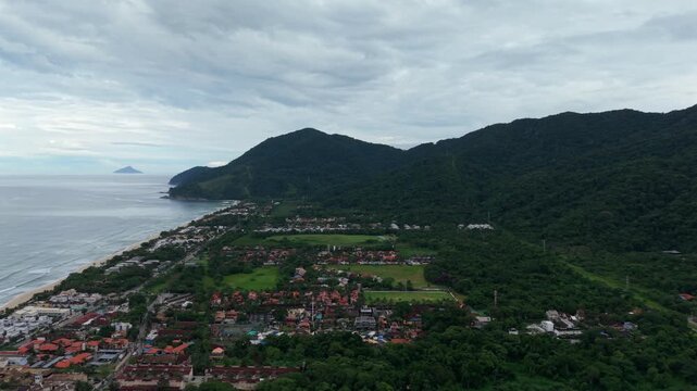 Maresias, S&atilde;o Sebasti&atilde;o, passing over the coastal town while approaching beach and ocean against backdrop of vegetated hills. Left tracking aerial.
