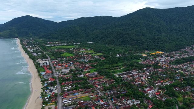 Coastal houses and rural buildings of Maresias, S&atilde;o Sebasti&atilde;o, set against lush green tropical mountains on cloudy tropical day. Forward aerial glide.