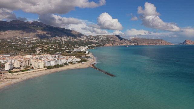 Albir and Altea Costa Blanca Spain.Backwards high drone flight over crystal clear blue sea of the beach and breakwater with views of Peacock Island, the mountains and interesting clouds in bright sun