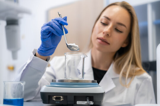 Laboratory technician carefully weighing precise amounts of chemical powder using a spoon over glass beaker on balance scale.
