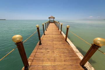 Naklejka premium Idyllic wooden pier with a thatched gazebo stretching into the calm turquoise sea under a clear blue sky. Serene summer vacation background, perfect for travel, relaxation, and tourism concepts.