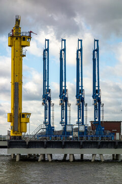 Industrial harbor scene featuring a yellow control tower and blue marine loading arms at an oil and gas terminal jetty, representing energy logistics and global trade.
