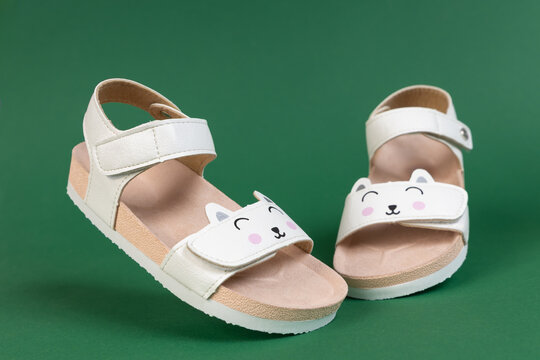 Pair of cute white children's sandals with cat face velcro strap on green background, adorable kids summer footwear product photography