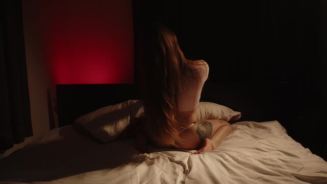 Back view of a seductive woman wearing enticing lingerie sensually thrusting while holding the headrest of a bed in dark bedroom