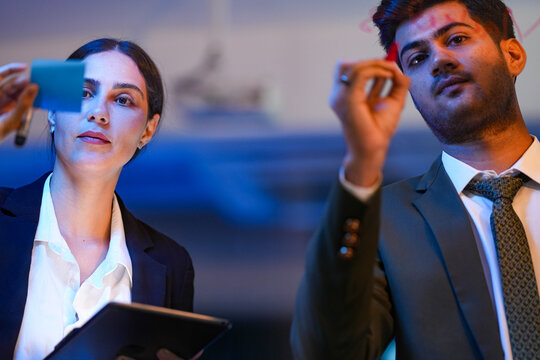 Diverse software engineers collaborating on generative AI algorithms, writing code on a glass board in a high-tech office at night. Modern tech innovation concept.