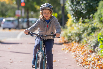 Naklejka premium Child rides bike on autumn. Kid wearing bike helmet. Happy child learning to bike ride. Child with bicycle in autumn fall park. Fall city scene with child on bike.