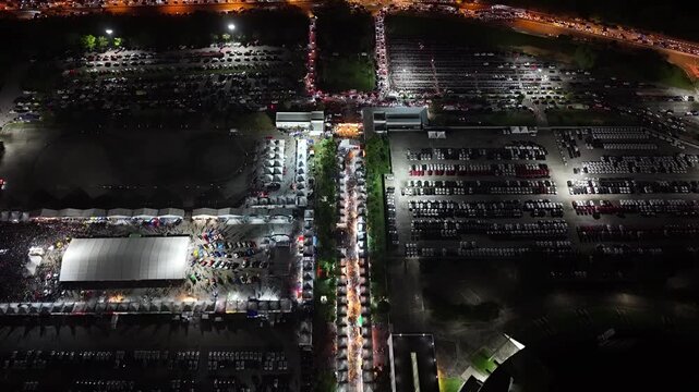 Drone aerial of Batu Kawan Modifighter Festival parking, tents, crowds and exhibit area at night.