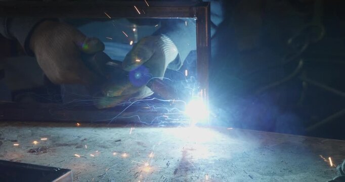 Industrial welder with protective gloves connecting metal profile pipes in a workshop, creating bright flashes of light, sparks, and smoke during the manufacturing process in a factor.