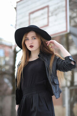 Fototapeta premium A girl walks down the city street in a leather waistcoat with a phone. Young beautiful girl in a hat and with a dark make-up outside. Girl in the Gothic style on street.