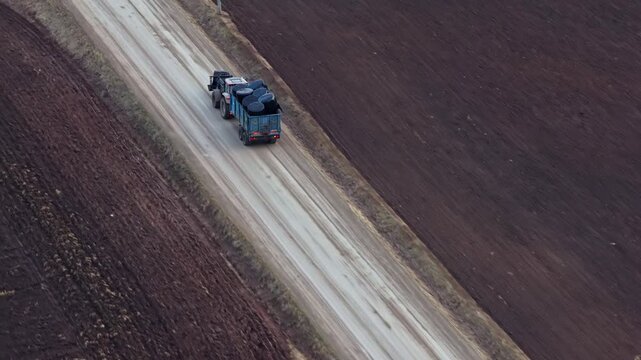 Tractor pulling a trailer with wrapped hay bales on a farm road, moving harvest in aerial follow shot
