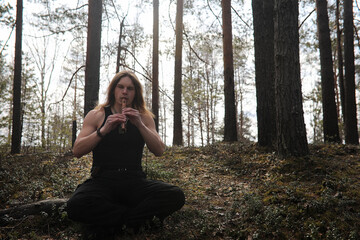 Obraz premium A musician with a tool in nature. A man is playing a flute in a pine forest. The music of the druids in the spring forest.