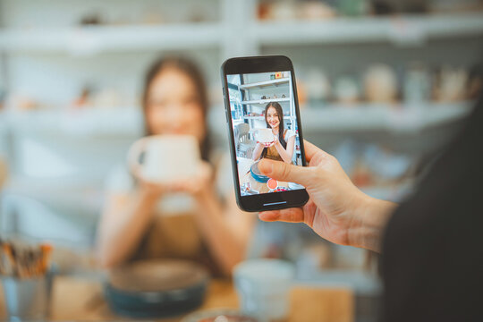 Close up of hand using smartphone to record happy young Asian woman holding handmade ceramic mug in pottery studio. Influencer creating social media content for DIY workshop. Bright, authentic