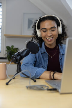 Non binary adult recording podcast at desk wearing headphones, denim jacket, using laptop and mic