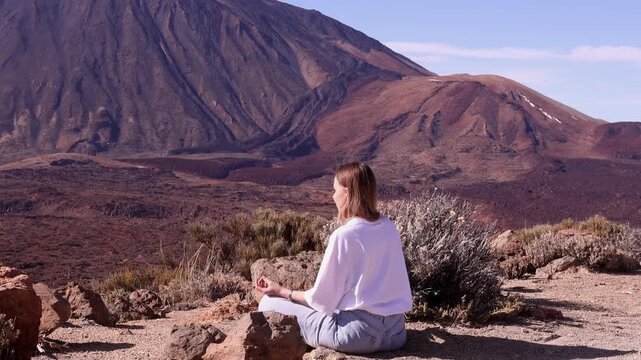 Young woman in white top sitting in lotus pose meditating on rocky ground with iconic Mount Teide volcano in background, Tenerife. Premium outdoor wellness and mindfulness travel content