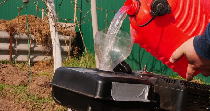Male farmer refueling a walk-behind tractor with gasoline from a red plastic jerrycan, using a makeshift funnel before starting agricultural work in the garden on a sunny spring day.