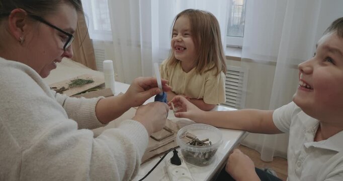 Happy family engaging in a creative hobby together, with the mother carefully showing her son and daughter how to use a hot glue gun for their art project at a table in a cozy room.