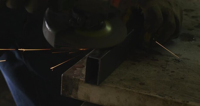 Skilled metalworker using an angle grinder to smooth the edge of a rectangular steel pipe, with hot sparks flying in a dimly lit industrial workshop before the welding process begins.