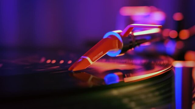 Orange phono cartridge playing vinyl record on turntable with neon purple and pink studio lighting.