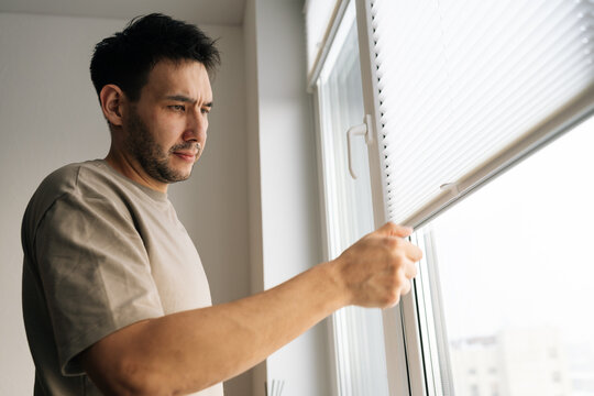 Thoughtful man with worried expression opens white pleated blinds looking outside. Natural light illuminates his face. Concept of waiting, anxiety, or curiosity.