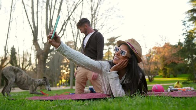 Happy woman wearing sunglasses and a hat taking a selfie with her phone while lying on a picnic blanket, as her boyfriend plays with their greyhound dog in the background on a sunny day