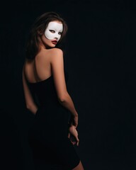 Fototapeta premium 白いマスクをつけた女性が黒いドレスを着て振り返っている姿。ファッションやミステリアスな雰囲気のポートレートに適した写真