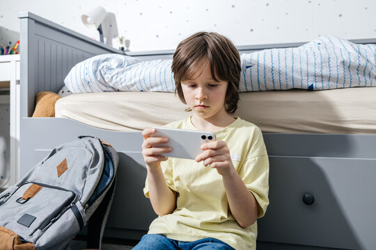 Student sitting in bedroom using smartphone near school backpack