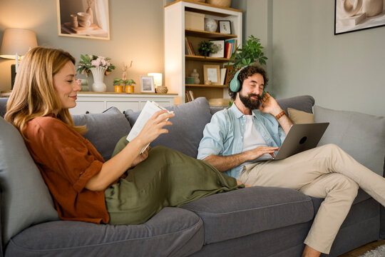 Couple relaxing on sofa reading and using laptop at home