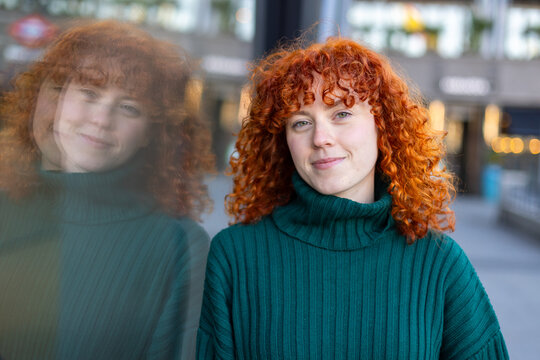 Smiling portrait of woman with red curly hair and reflection in city glass