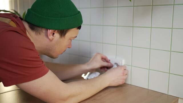 Young male electrician carefully installing a new power outlet on a tiled kitchen backsplash, working on the electrical wiring with a screwdriver during a home renovation project