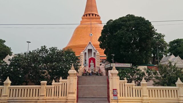 Camera moves in reverse along paved walkway revealing wide entrance leading toward giant orange stupa while hazy morning light illuminates green trees surrounding sacred Buddhist site in Thailand