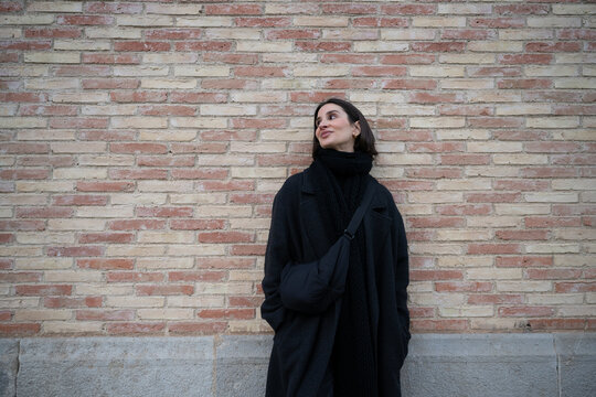 Fashionable woman wearing a black coat poses confidently against a brick wall backdrop in an outdoor setting