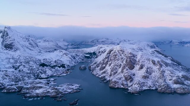 Breathtaking drone shot of snowcovered peaks and dark blue fjords in Lofoten, Norway during winter twilight. Ideal for travel documentaries and nature wellness content.