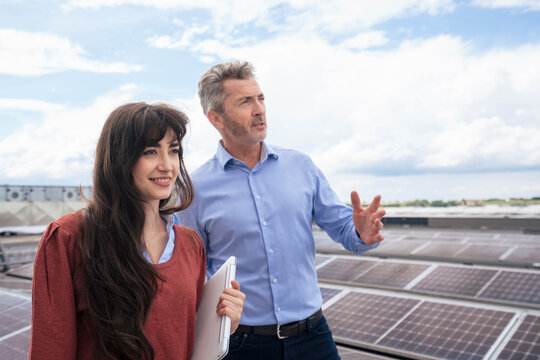Young businesswoman holding laptop discussing with mature colleague on rooftop under cloudy sky