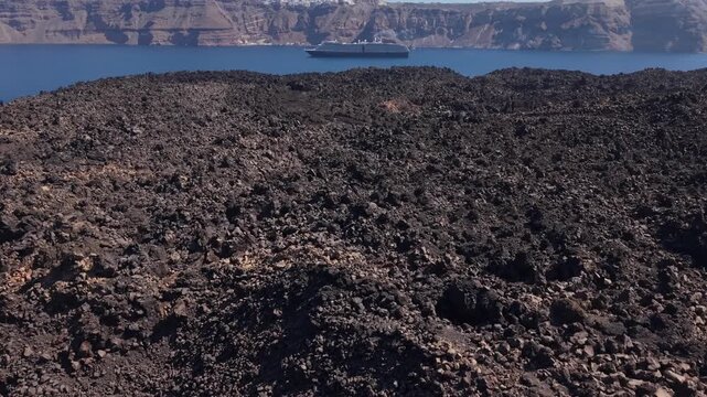 Drone flying over lava fields at Santorini volcano revealing layered volcanic terrain, the caldera, and a cruise ship in the Aegean Sea.