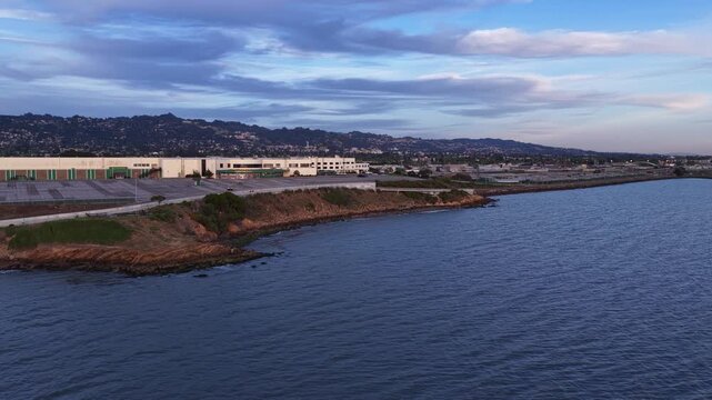 Aerial drone view moving forward over the san francisco bay towards the albany bulb, with berkeley hills in the background under a cloudy sky during a beautiful california sunset