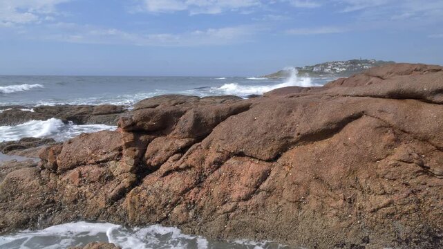 Crabs scurry in and out of rocky crevices as waves crash against weathered red- brown rocks on a sunny day, with a small coastal town visible on a distant green hill under a blue sky with clouds.