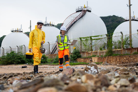 Environmental engineers walking through rocky terrain near large fuel storage tanks, Technical team in protective gear carrying tools and laptop performing a gas refinery industrial site inspection