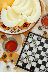 Checkers game board with hand holding piece and sliced fresh yellow melon on floral tablecloth in warm summer sunlight top view © Юрий Ерохин
