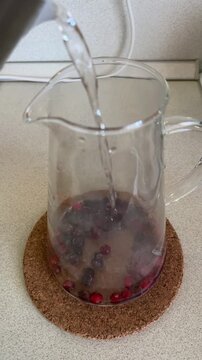 Glass pitcher with frozen currants and cranberries, boiling water poured, steam rising on tempered glass, berries releasing color