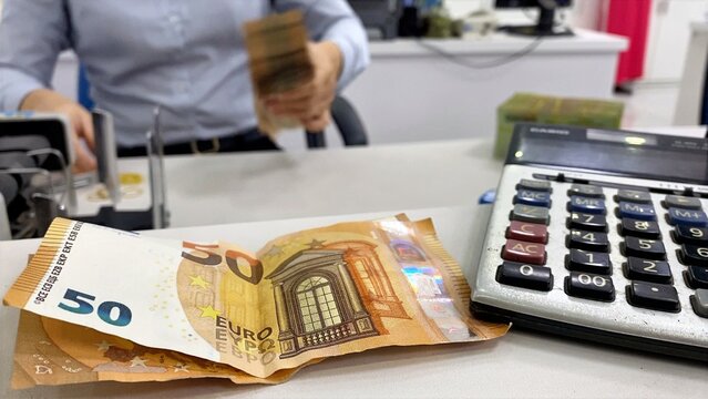 Person handling cash and using a money counter, with euro banknotes and a calculator resting on the counter, showing financial transactions and banking operations