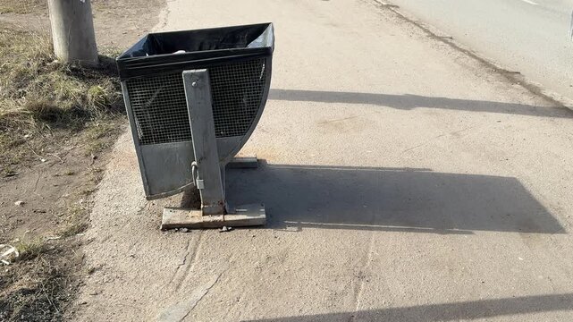 Daytime roadside dumpster with black bag, sunlit asphalt, metal bin casting long shadow near lamppost, gravel verge and curb
