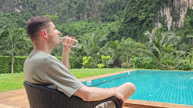 Man sitting in chair drinking water from glass bottle near swimming pool with tropical plants and limestone cliffs in background relaxing outdoor travel and vacation lifestyle scene