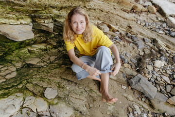 Fototapeta premium Woman sitting on rocky seashore and rolling up jeans while looking at camera
