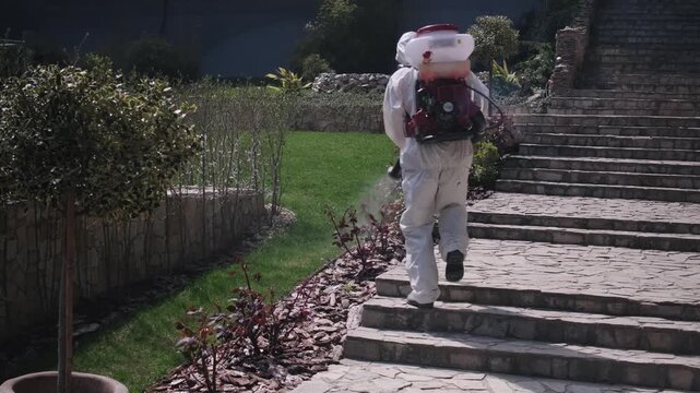 Worker in protective suit spraying garden with pesticide using backpack fogger, outdoor pest control treatment in residential yard